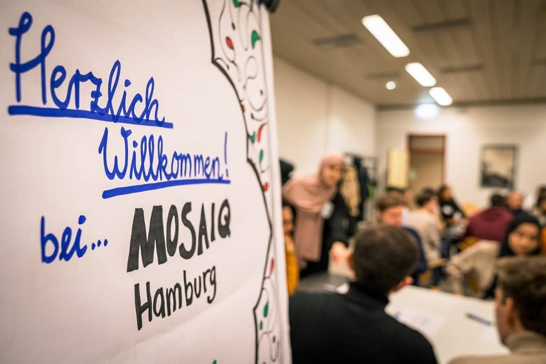 Auf der linken Seite ist ein Flipchart-Papier zu sehen, auf welchem geschmückt "Herzlich Willkommen bei Mosaiq Hamburg" steht. Auf der Rechten Seite ist ein Raum zu sehen, in verschwommener Optik, auf dem mehrere Personen miteinander reden.  
